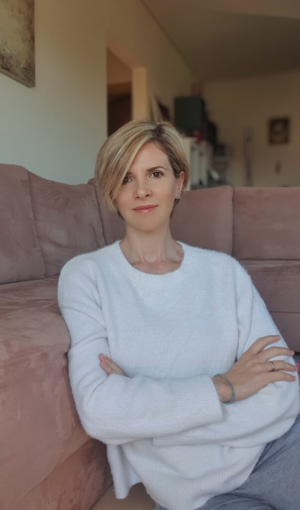 Γυναίκα ξανθιά, με λευκή μπλούζα, καθισμένη με σταυρωμένα τα χέρια στους ώμους, διπλα σε καναπε μπεζ χρωματος